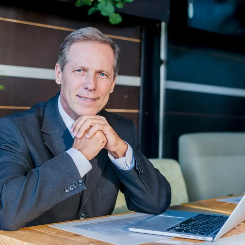 Homem de meia idade com cabelo branco e usando terno sentado em uma mesa com o notebook aberto. Ambiente bem iluminado e com algumas plantas em composição na imagem