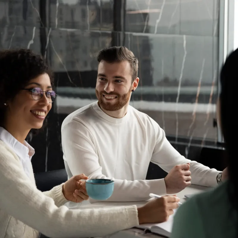 Homem jovem de blusa de manga comprida branca, sorrindo e olhando para uma mulher jovem de cabelos cacheados, blusa de manga comprida branca e sorrindo. Ambos conversando com uma outra mulher de costas em um ambiente comercial de trabalho