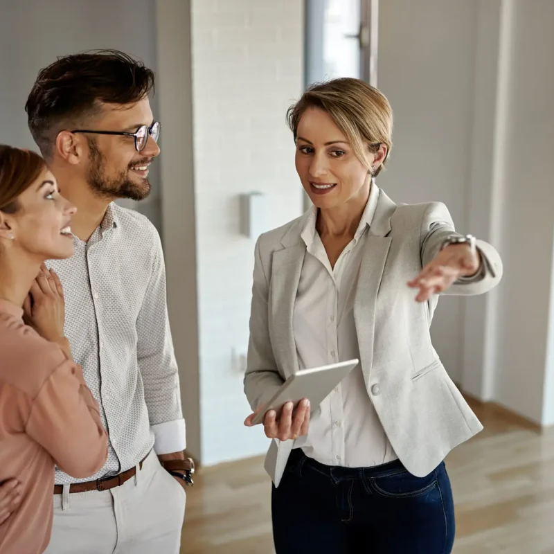 Um casal jovem, sorridente, conversando com uma corretora de imóveis em um ambiente moderno e iluminado. A corretora, segurando um tablet, gesticula enquanto apresenta o espaço ao casal.