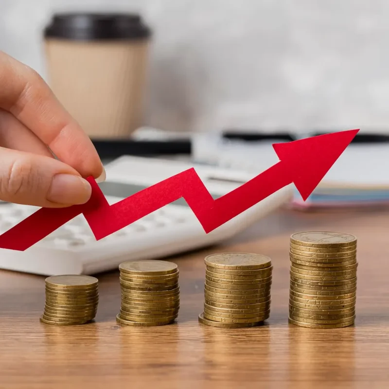 Mão segurando uma seta vermelha em cima de pilhas de moedas com ordem crescente. Fundo da imagem é uma mesa com calculadora, papéis e copo de café