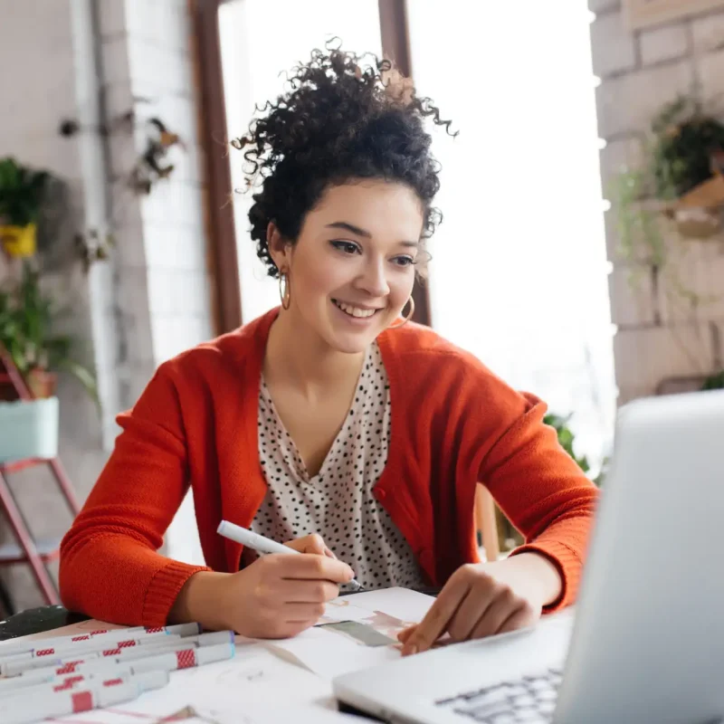Young smilng woman with dark curly hair sitting at the table happily working on laptop drawing fashion illustrations spending time in modern cozy workshop with big windows