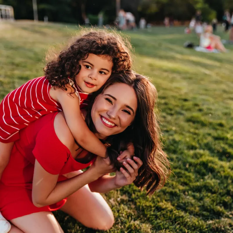 Refined little girl embracing sister on nature background. Happy female model with brown hair playing with curly kid in park.